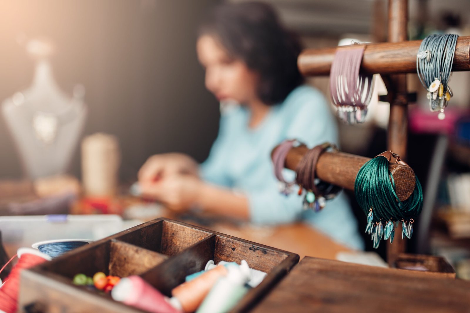 Atölyede el yapımı takı üreten bir zanaatkâr ve ön planda sergilenen el yapımı bileklikler ve aksesuar malzemeleri