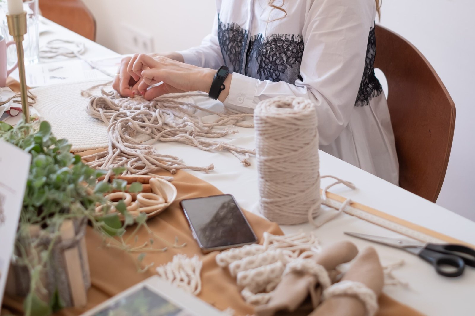 Bir kadının masada makrome düğümleme çalışması yaparken, ipler, makas ve doğal malzemelerin bulunduğu el işi çalışma ortamı.